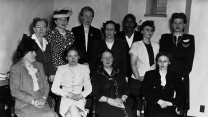Eleven women in mid 20th century business attire stoically pose for a photo. Four are seated in front. Seven stand behind them. They are all middle-aged to elderly in age. And most are Caucasian, minus one or two who may be African American.