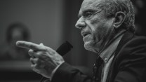 A black-and-white photo of Robert F. Kennedy Jr. in profile, talking into a microphone and pointing with the index finger of his left hand.