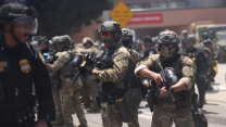 Lines of masked agents dressed in green military fatigues and battle helmets hold weapons as they prepare to confront protestors.