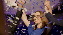 A triumphant Sarah McBride smiles amid a flurry of confetti, as she holds the raised hands of supporters above her head. McBride is wearing glasses and a blue dress.
