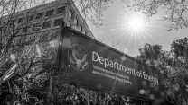 A black and white, low-angle shot of a sign for the Department of Energy. The sign also includes the address "1000 Independence Avenue SW, James V. Forrestal Building" and the department's seal. In the background, the sun shines brightly from behind the corner of a large, modern office building, with tree branches framing the top and sides of the image.