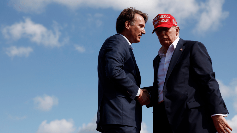 Glenn Youngkin and Donald Trump, both in blue suits without a tie, shake hands outdoors under a partially cloudy blue sky. Youngkin appears to be speaking to Trump, as Trump, wearing a MAGA baseball cap, leans in to listen.