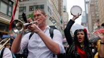 A man in a white shirt and glasses plays a trumpet while marching through the streets of Manhattan. Beside him, a woman dressed in black with a red flower in her hair raises a tambourine in the air, singing or chanting. Behind them, a large crowd and urban buildings are visible, suggesting a festive protest atmosphere.