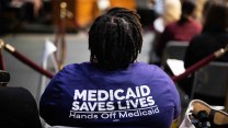 A black woman wearing a shirt that says on the back "Medicaid Saves Lives: Hands off Medicaid" at a hearing.