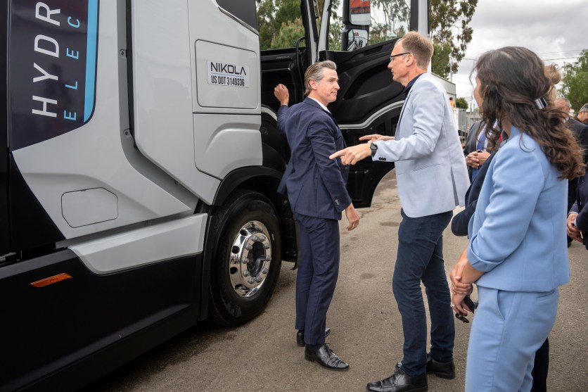 Closeup image of a Nikola Truck with the passenger door slightly ajar and Gov. Gavin Newsom in a suit standing in front of it alongside two other people.