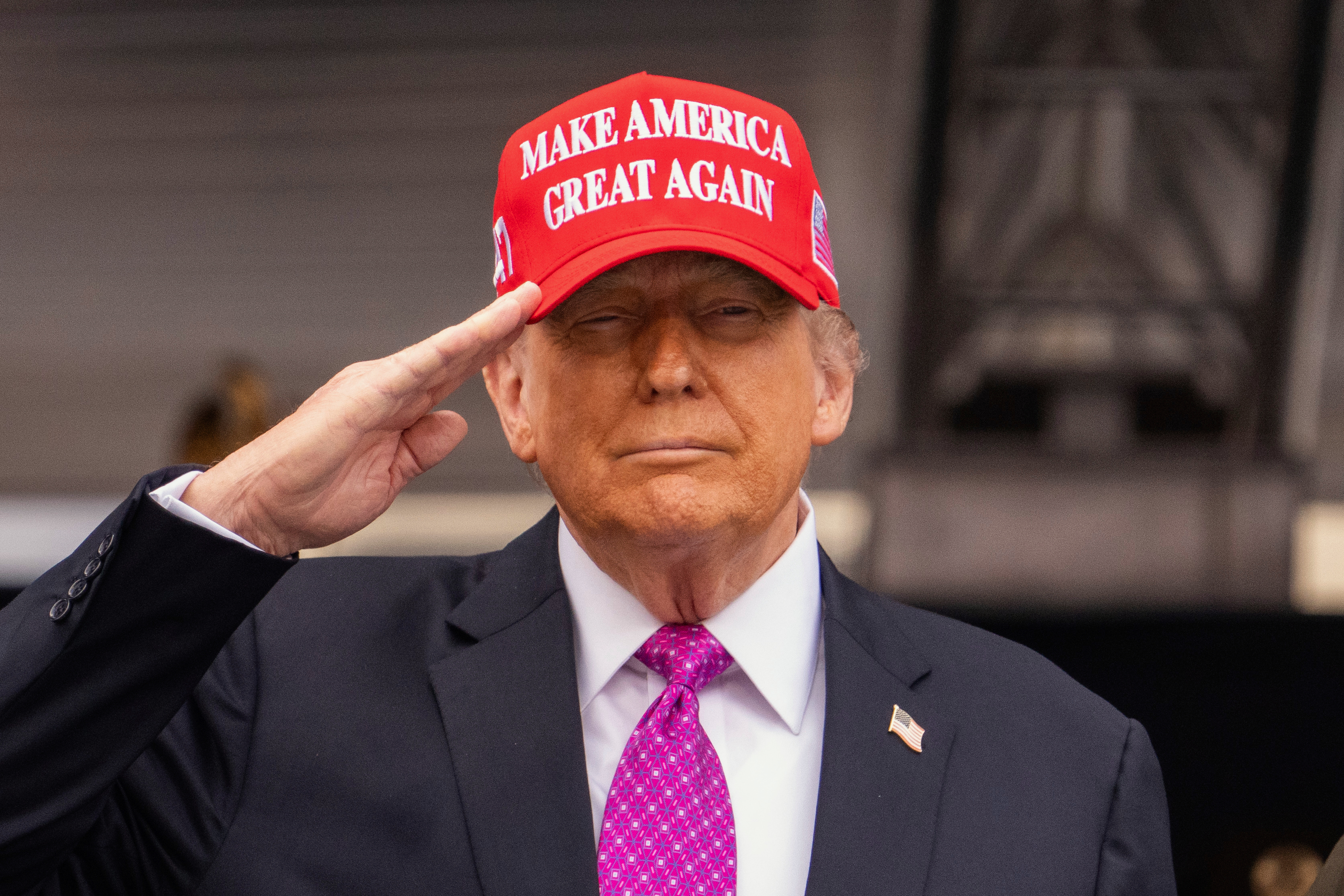 A close-up photo of Donald Trump saluting. He wears a dark suit, pink patterned tie, and red "Make America Great Again" baseball hat.