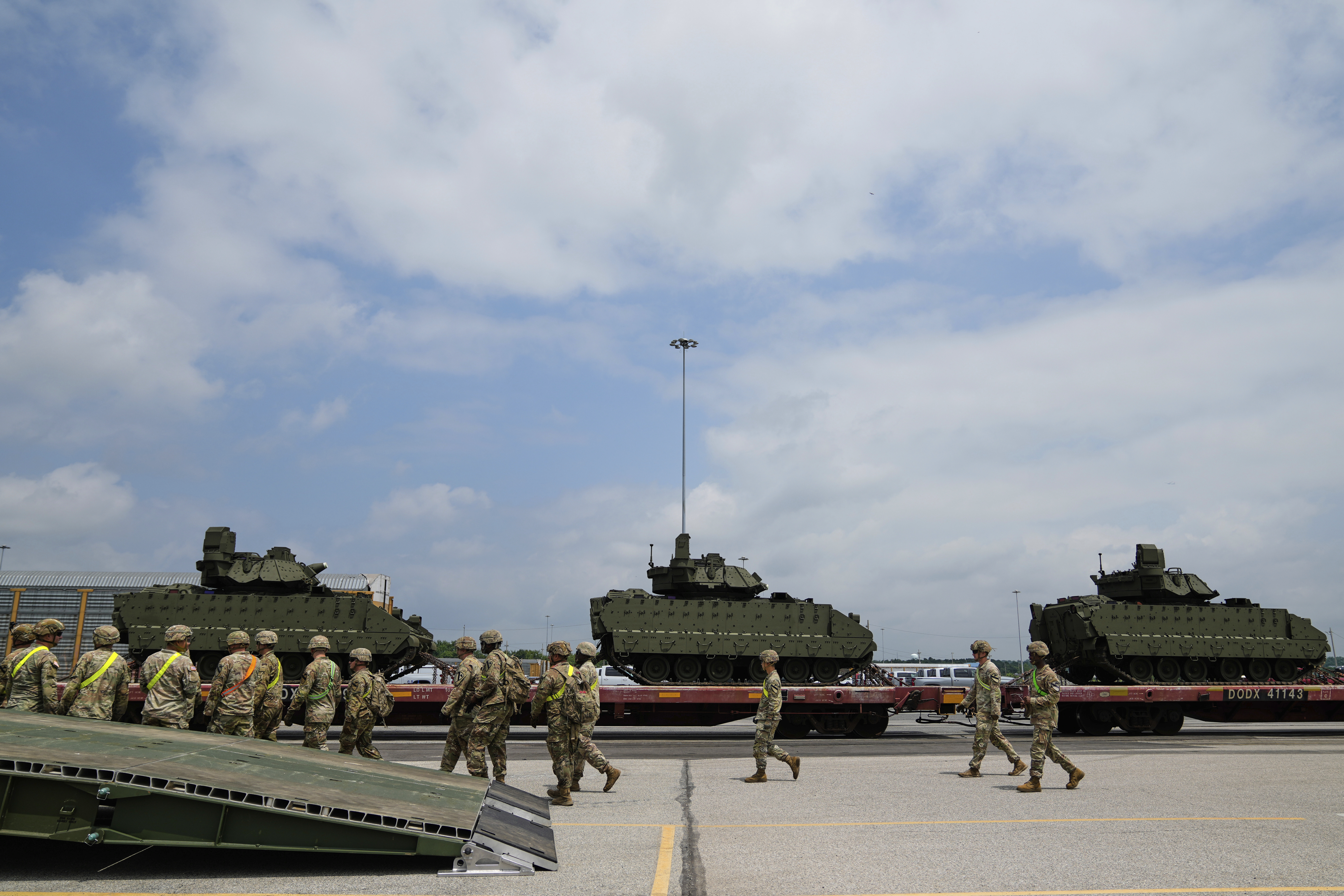 Troops in camouflage uniforms and helmets walk past low flatbeds carrying tanks.
