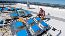 a white man kneels on a white roof next to a bunch of strange devices with blue squares on top, measuring heatheat