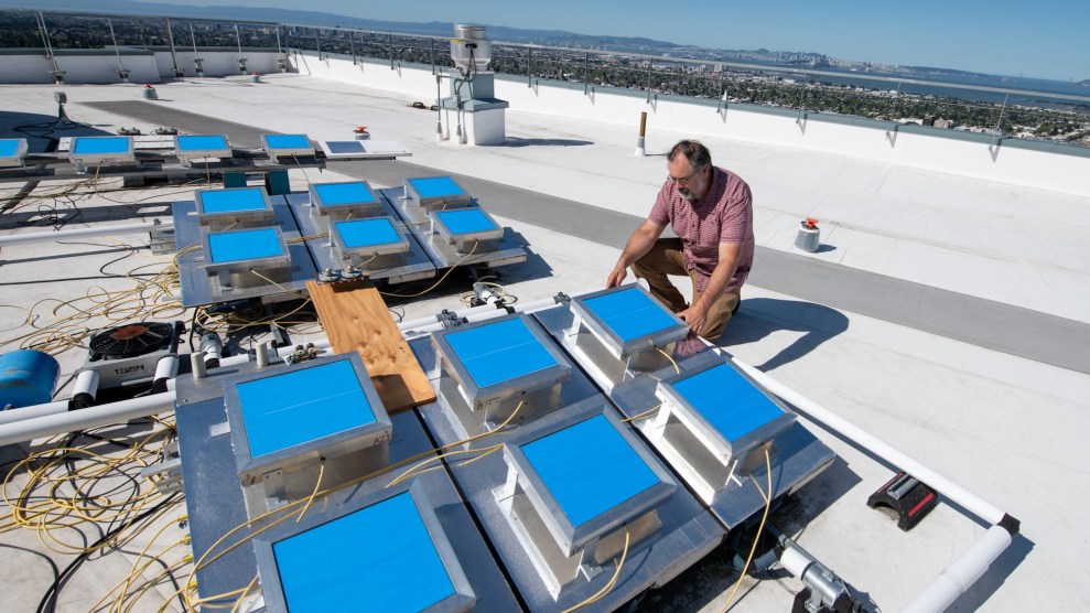 a white man kneels on a white roof next to a bunch of strange devices with blue squares on top, measuring heatheat