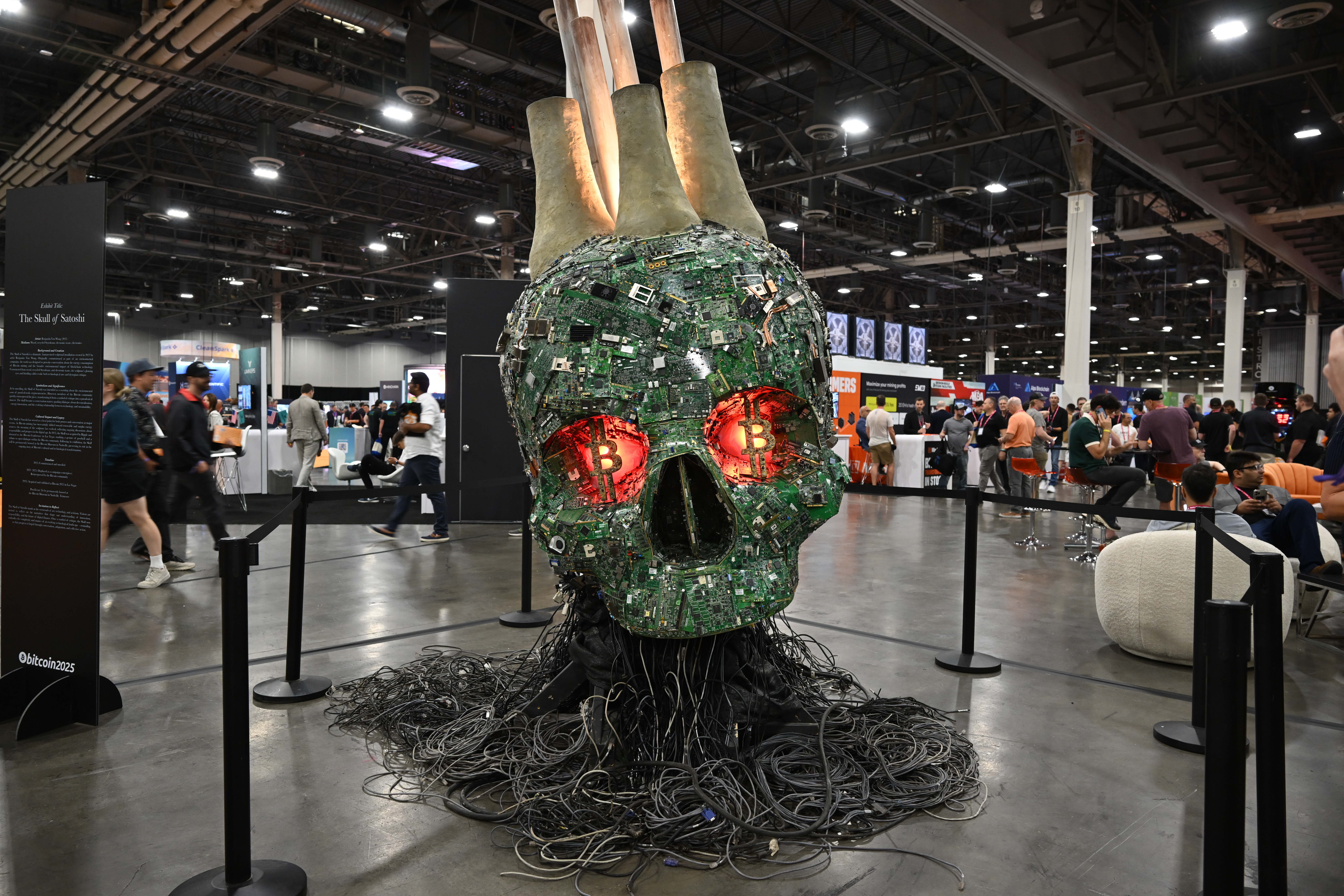 Art instillation of a large skull made of circuit boards and computer servers with glowing bitcoin symbols for eyes