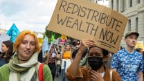 One person in the center of the image holds a sign that reads: redistribute wealth now. Two others are in the foreground, a group of people behind them are hold signs.