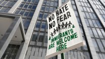 A white protests sign against the backdrop of a tall official building, steel and glass, reads "no hate, no fear, immigrants are welcome here."