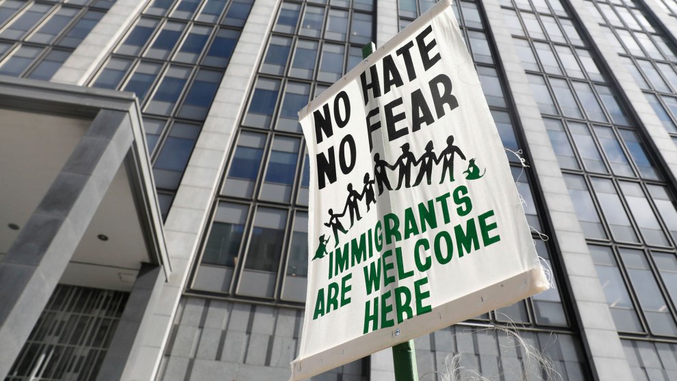 A white protests sign against the backdrop of a tall official building, steel and glass, reads "no hate, no fear, immigrants are welcome here."