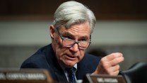 Sheldon Whitehouse, wearing a blue jacket and tie, gestures with two fingers as he speaks. He is sitting behind a desk, his name card out of focus.