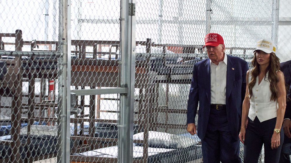 A tired-looking president Trump in blue suit, no tie, trudges with homeland security sec Kristi Noem in white shirt and ball cap past a indoor cage filled with bunk beds.