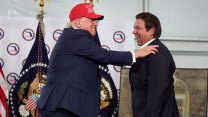 An overweight donald trump in a blue suit wearing a maga hat grins as he lays a hand on the shoulder of a guffawing Rick Desantis in a dark suit on a makeshift stage with blue presidential flags.