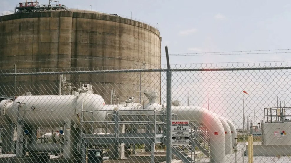 A gas plant as seen through a chain link fince, with large white pipes and tanks and a much larger cylindrical concrete structure looming in the background
