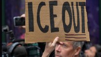 A hand holds a cardboard sign with "ICE OUT" written in black sharpie. Another man's face, a middle-aged white guy, can be seen in the background, but it looks like someone else's sign. Others can be seen recording with smartphones.