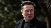 Elon Musk is wearing a black coat and making a puckered facial expression, looking to his left, with a blurred outdoor background.