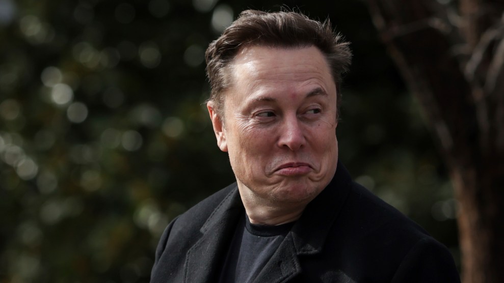Elon Musk is wearing a black coat and making a puckered facial expression, looking to his left, with a blurred outdoor background.