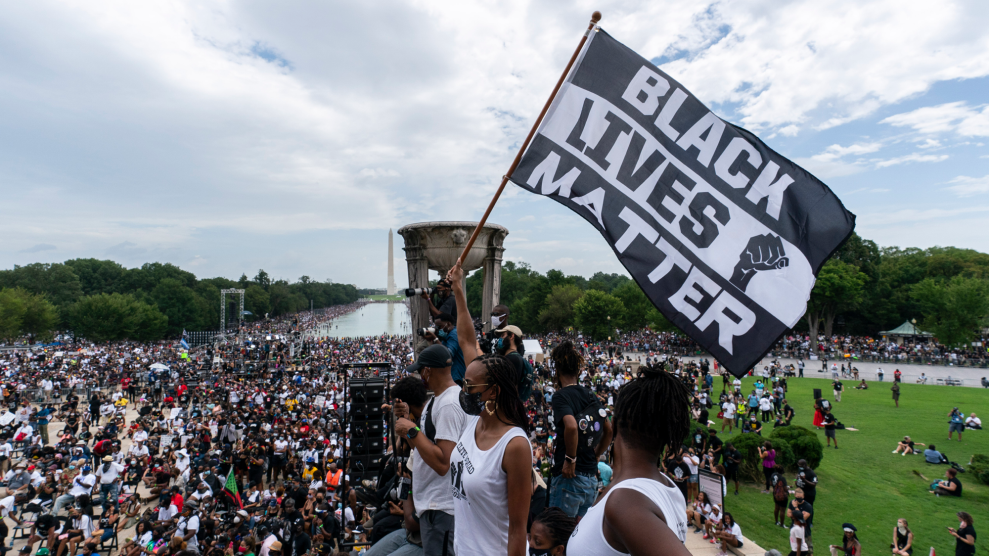 A woman extends her right arm above her head as she holds a pole with a billowing "Black Lives Matter" flag. Standing on the steps of the Lincoln Memorial in Washington, D.C., the woman, dressed in a white tank top, white shorts and a black mask, is among thousands gathered around the reflecting pool.