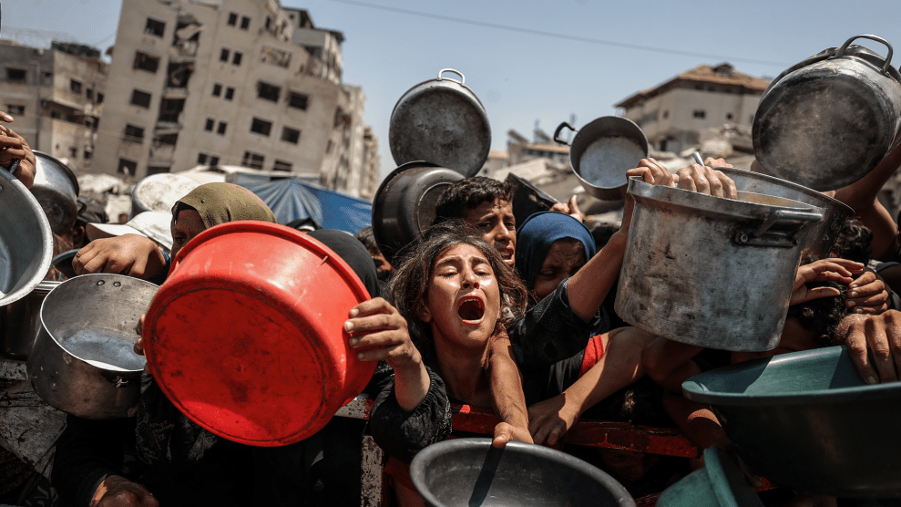 People, mostly young children, wail and clamor as they lean against a barricade while holding empty pots and buckets. Behind them are the ruins of bombed buildings against a blue sky.
