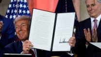 Smiling Donald Trump holds up a signed executive order, his signature huge, while a man claps in the background