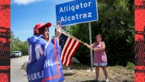 Photo collage of a young woman wearing a "Make America Great Again" hat and a "TRUMP" banner draped around her shoulders, taking photos in front of the sign for Alligator Alcatraz. A woman in the background smiles and waves a 1776 American flag. To the left and right of the photo is a textured and stylized scene of chainlink fence cells in a detention center.