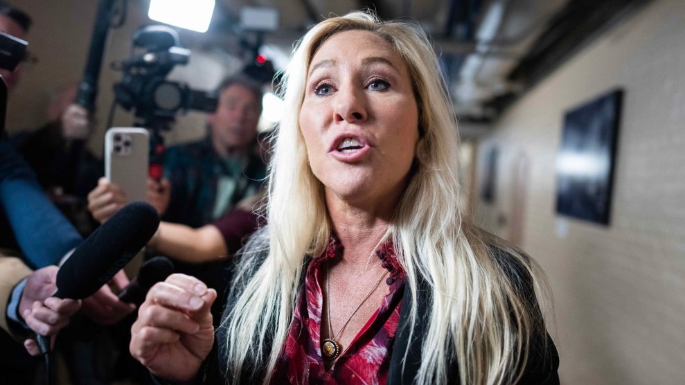 Rep. Marjorie Taylor Greene speaking to a throng of reporters holding cameras, microphones, and lights in a hallway somewhere.