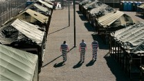 Three incarcerated men in black-and-white-striped uniforms walk through the courtyard in an outdoor jail composed of tents.