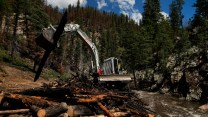 A large machine operated by a person sits in the middle of a river, with a pile of tree debris to the side of it, next to the river. There are pines in the background against a blue sky.