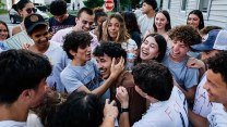 A smiling boy in a brown shirt is surrounded and hugged by a group of people wearing white and grey shirts.