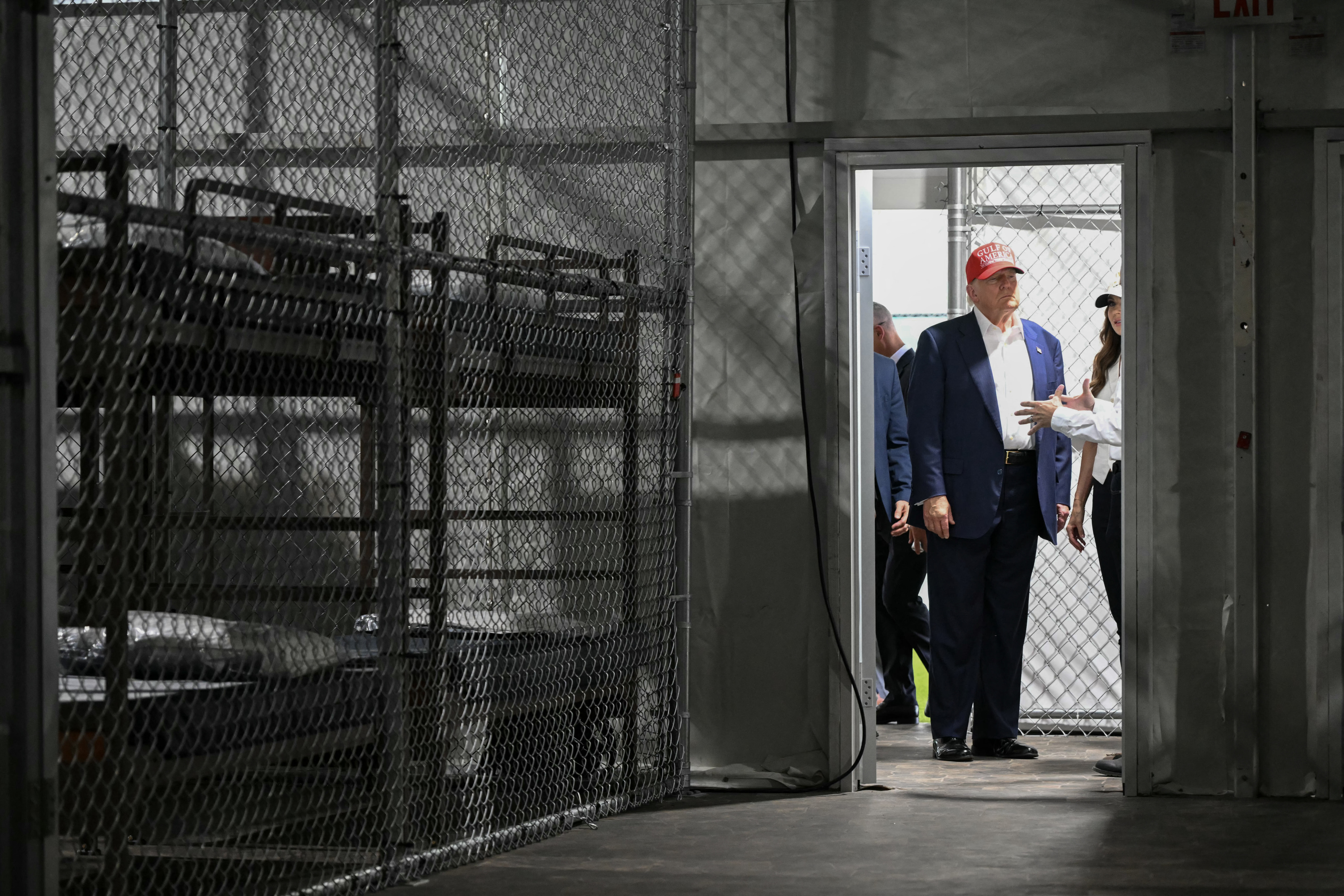 President Trump looks in on a migrant detention center, towards bunk beds behind caged walls.
