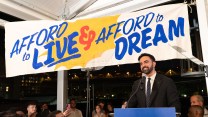 Zohran Mamdani wearing a black suit and tie stands in front of a banner that says "Afford to live and afford to dream."