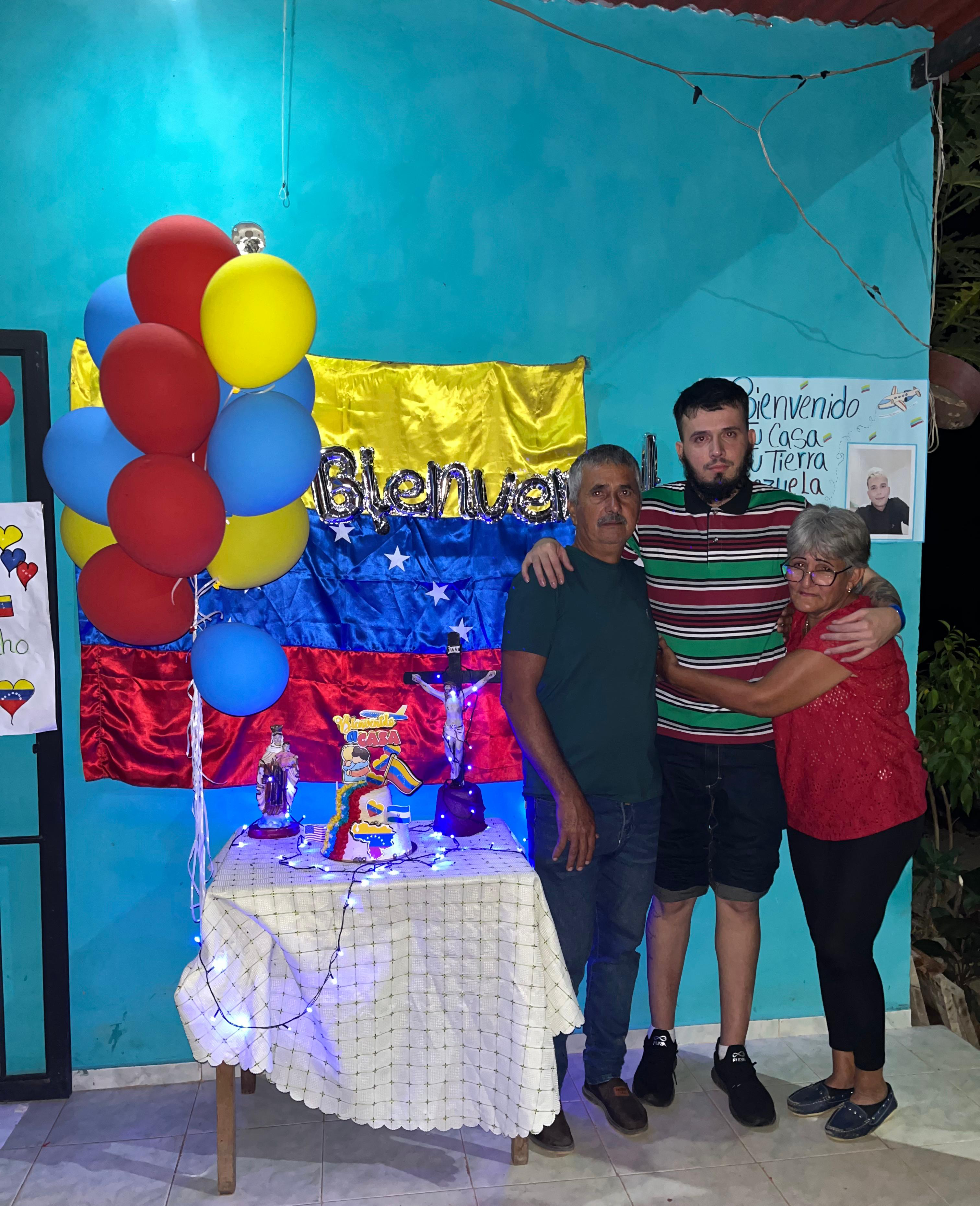 A young bearded man stands with his arms around a middle-aged man on the right and a woman on the left. Beside them are the Venezuelan flag and balloons in the same color as the flag: red, blue and yellow.