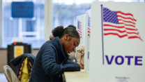 A young African American woman wearing a dark blue coat sits in a folding chair as she fills out her ballot on a table. Erected on the table are several white partitions that have a billowing American flag design on them with the word “Vote” underneath.