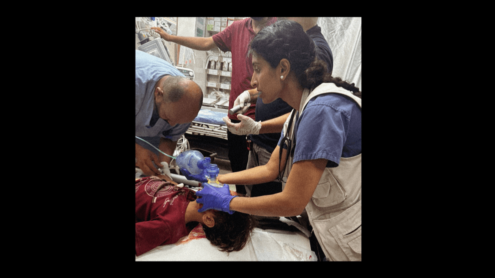 A woman wearing blue medical gloves, a tan vest, and a dark blue short-sleeve scrub top places a breathing apparatus over the face of a child in a maroon T-shirt lying in front of her. A middle-aged man assists her.