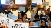 A crowd of women with signs, mostly saying "put Texans first," stand on a stairway at the Texas statehouse and cheers returning democratic politicians.