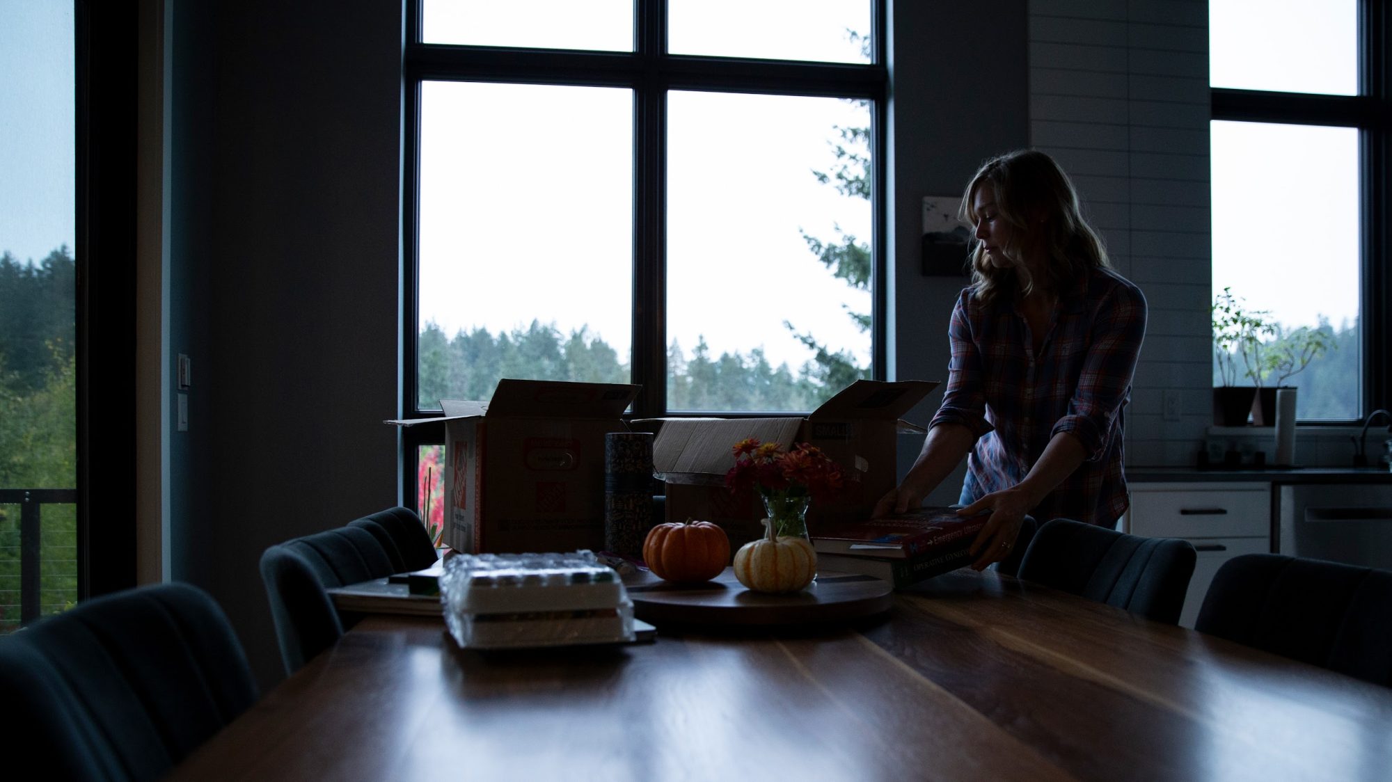 A woman unpacks boxes in the kitchen of a house with a view of evergreen trees.