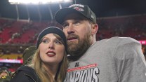 Taylor Swift and Travis Kelce pose for a photograph in a football stadium. She wears a black beanie and a black sweatshirt. He wears a Kansas City Chiefs cap and a grey t-shirt that says “Champions”