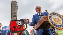 A white man holds a comically large chainsaw with the words "Cuts to our future" on it.