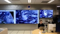 A person sits in front of a laptop in front of large screens with hurricanes displayed on them.
