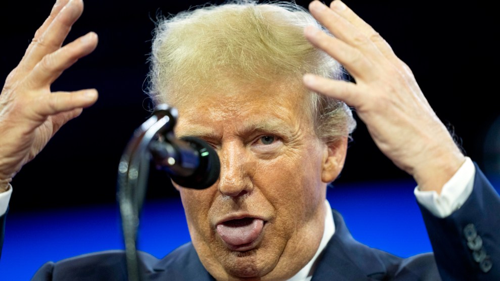 A person in a dark suit and red tie, Donald Trump, stands behind a microphone, raising both hands with palms outward while sticking out his tongue.