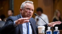 Robert F. Kennedy sits at a microphone with both hands in the air, palms down, as he talks