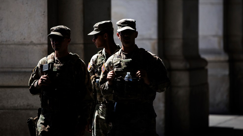 Three National Guard troops in camouflage uniforms stand in a cluster under the shade of a building. Their faces are obscured in shadow and in sharp contrast to the sunlight.