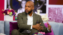 A middle-aged African American man sits on a blue sofa with square purple pillows. The man has a shaved head and a black beard that’s beginning to gray at the chin. He’s wearing an olive-colored suit and white shirt without a tie.