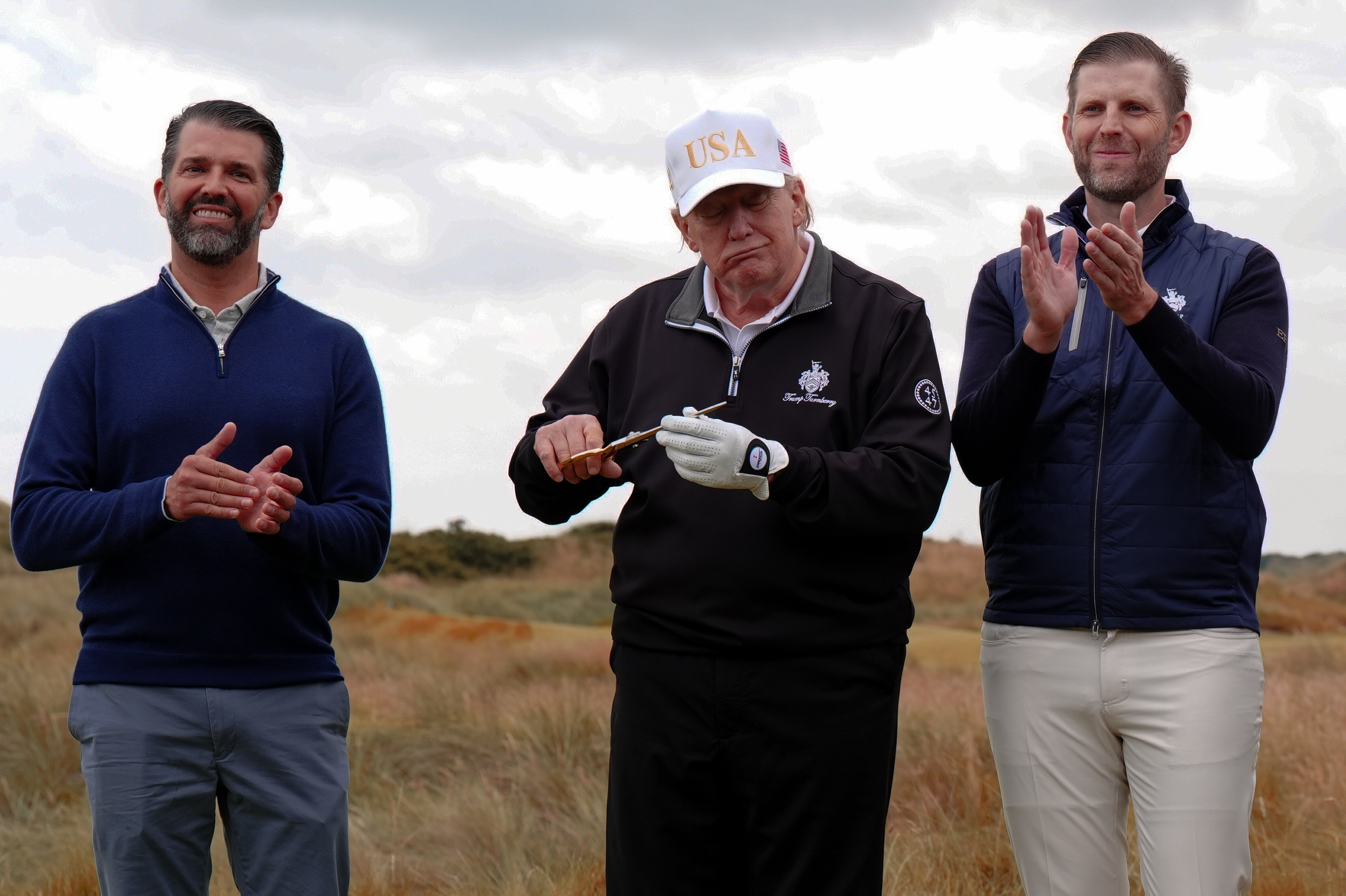 Donald Trump, dressed in a quarter-zip jacket and white USA baseball cap, holds golden scissors ceremoniously on a golf course with long grass behind him. His bearded sons flank him, clapping.