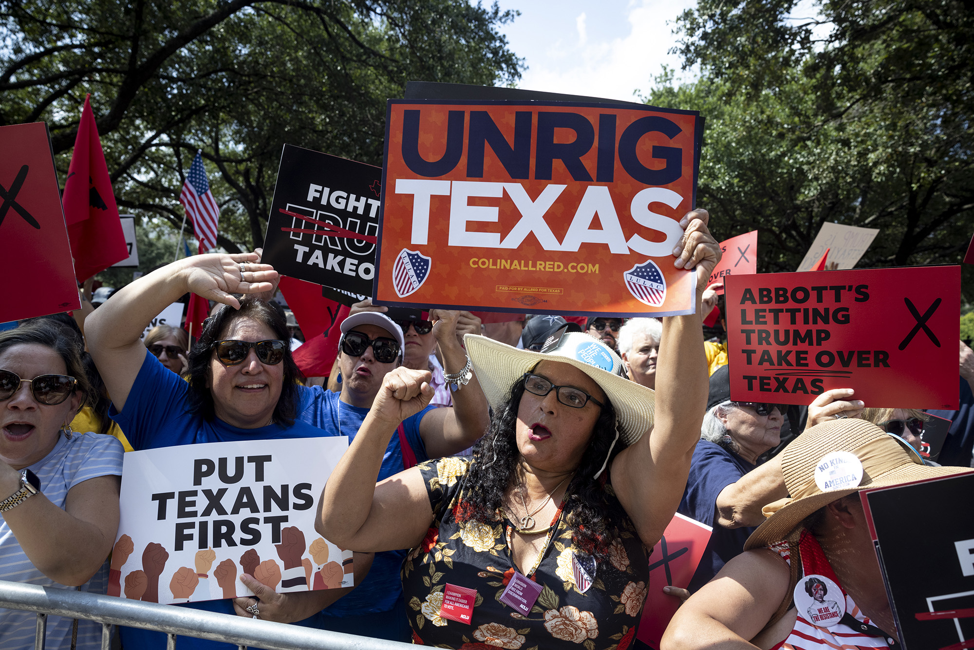 People in a crowd at a rally hold signs that read, "Unrig Texas," "Put Texans First," and "Abbott's letting Trump take over Texas."