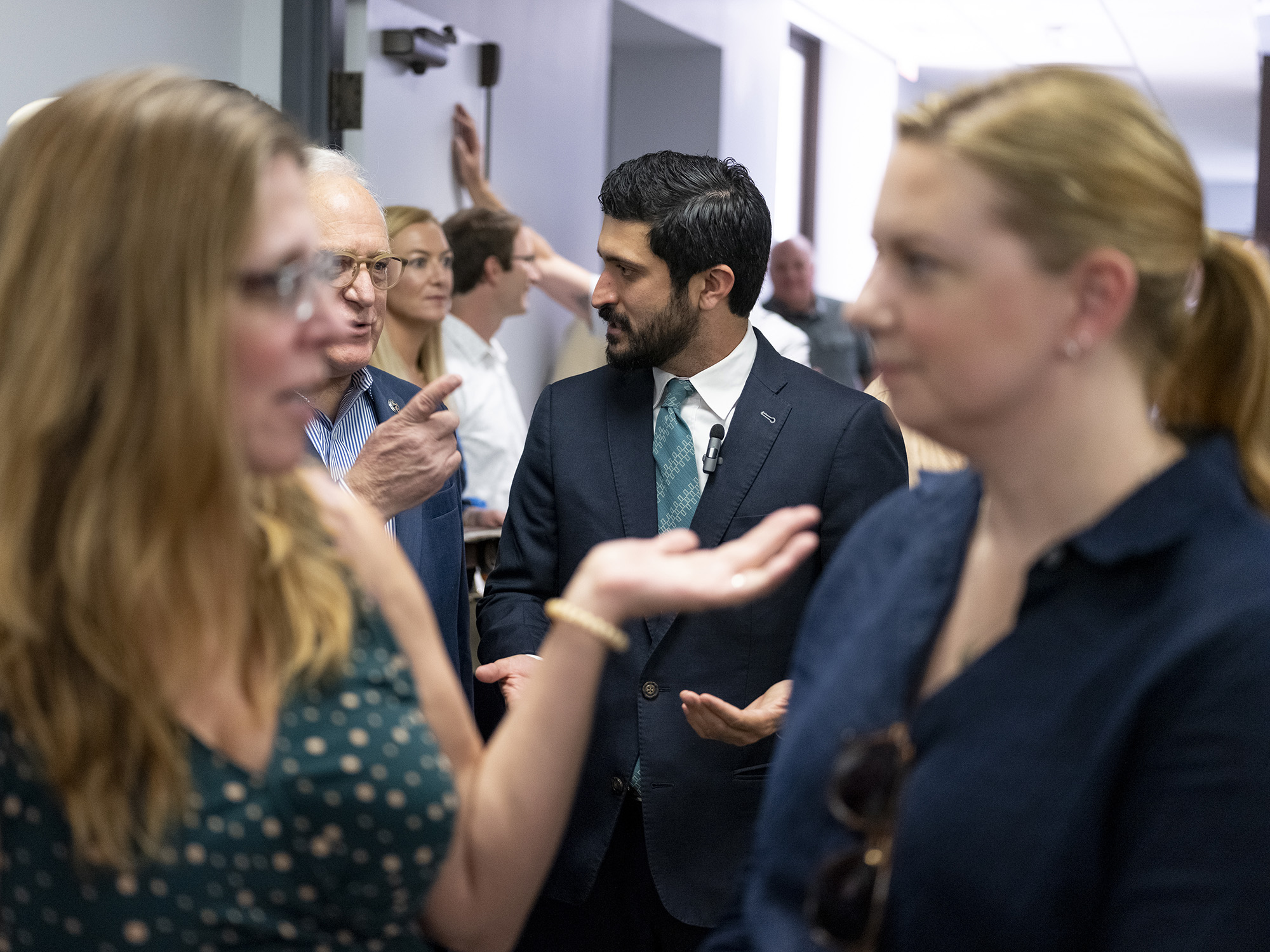 Shown behind two women in the foreground, who are out of focus, Greg Casar, dressed in a suit and tie, talks to a man.
