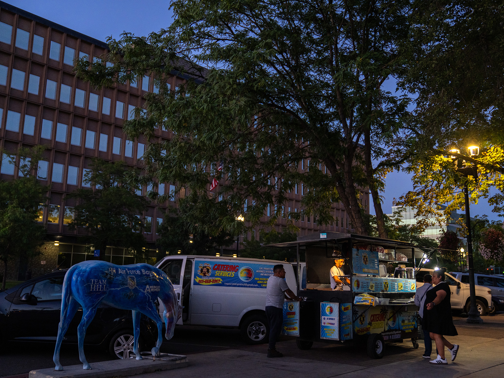 A street food cart near a painted statue of a horse at dusk, with an office building in the background.